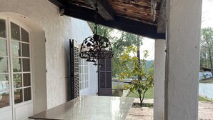 A covered patio area with a stone-tiled floor, a white column, and a hanging metal lantern. - Large villa with pool (Saint-Paul-En-Forêt)