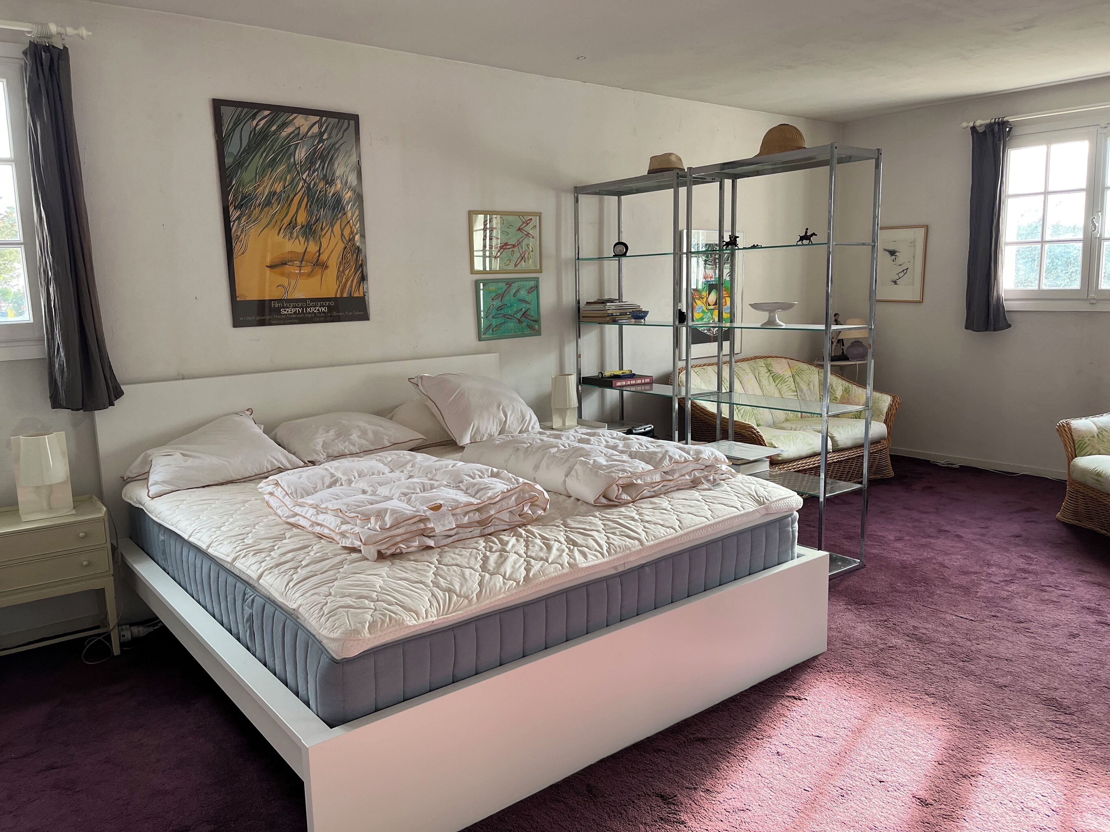 A bedroom with a bed, a glass shelving unit, and artwork on the walls.