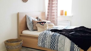 Ein Schlafzimmer mit Holz-Kopfstück, einem Bett mit gemusterten Betttextilien, einer schwarzen Tagesdecke, einem gewebten Korb und einem Fenster mit Vorhängen.