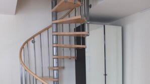 Moderni, spiraalimainen portaikko, jossa on puiset askelmat ja metallikaiteet, sijaitsee huoneessa, jossa on puulattia ja valkoinen seinä.
