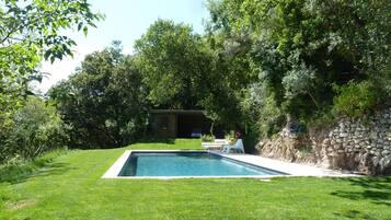 Une piscine rectangulaire entourée d’une pelouse bien entretenue, avec un mur de pierre et une petite construction en bois en arrière-plan.