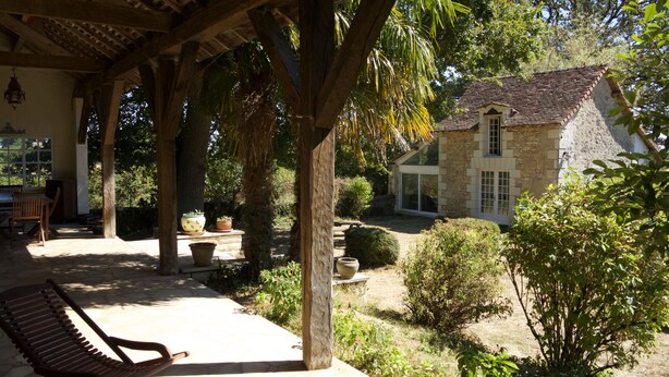 Une terrasse couverte avec un banc en bois, une maison en pierre avec un toit en tuiles et une végétation luxuriante tout autour.