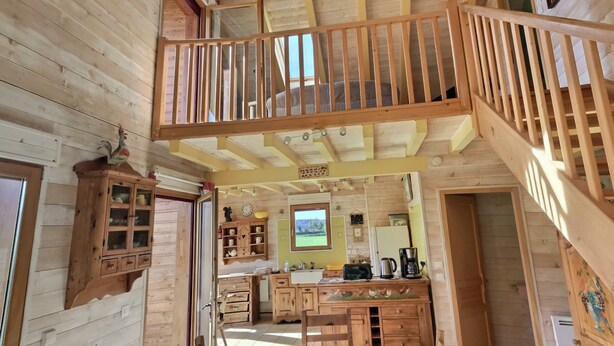 A wooden cabin interior with a kitchen area, wooden staircase, and a balcony with a railing.