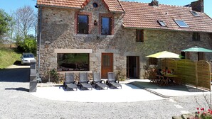 A stone house with a tiled roof, a front porch with chairs, and a table with umbrellas. - Gite "Le Bocage" - sleeps 6 - rated 2 stars (SARTILLY BAIE BOCAGE)