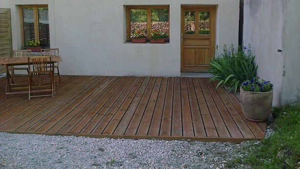 Una terraza de madera con una mesa y sillas, un pequeño jardín con flores y una puerta con ventanas.