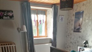 Un dormitorio con una cama, una cuna, una silla roja, un radiador y una ventana con cortinas.
