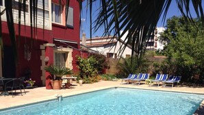 Una piscina con sillones de descanso, un edificio rojo y un jardín con plantas en macetas.