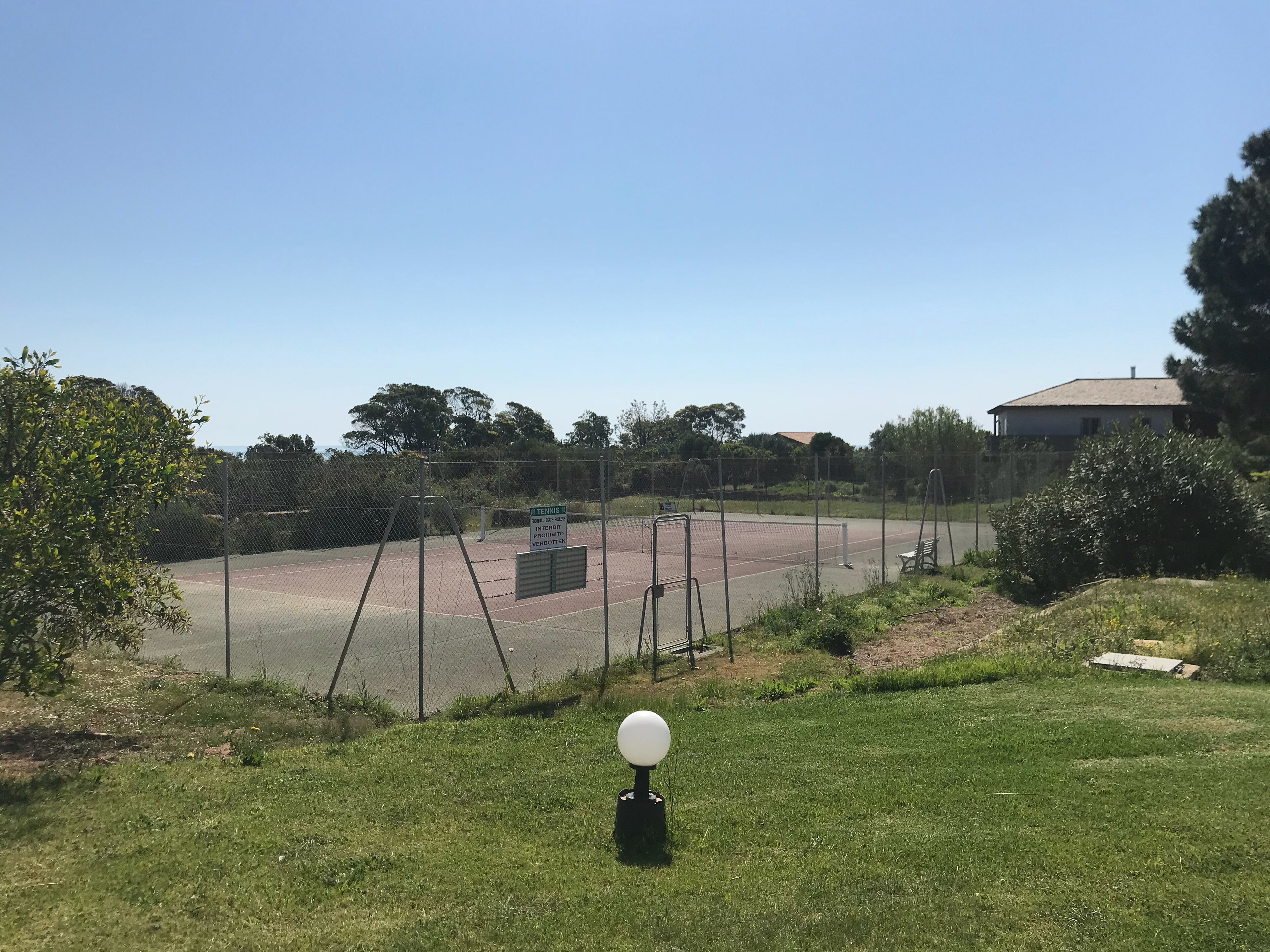 Una cancha de tenis con red, rodeada por una cerca y un objeto esférico blanco en el suelo.