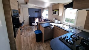 A compact kitchen with a stove, sink, and microwave. There is a small dining area with a table and chairs. - New mobile home (6/8 pers.), All options, Camping Marestang 4 * Location 23 (Canet-en-Roussillon)