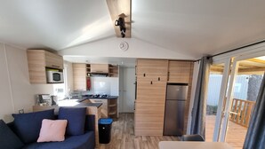 A compact living space with a kitchenette, a sofa, a dining table, and a view of the outside deck. - New mobile home (6/8 pers.), All options, Camping Marestang 4 * Location 23 (Canet-en-Roussillon)