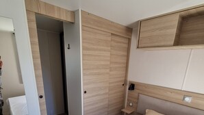 A modern bedroom with a wooden wardrobe, a bed with white bedding, and a small bedside table. - New mobile home (6/8 pers.), All options, Camping Marestang 4 * Location 23 (Canet-en-Roussillon)