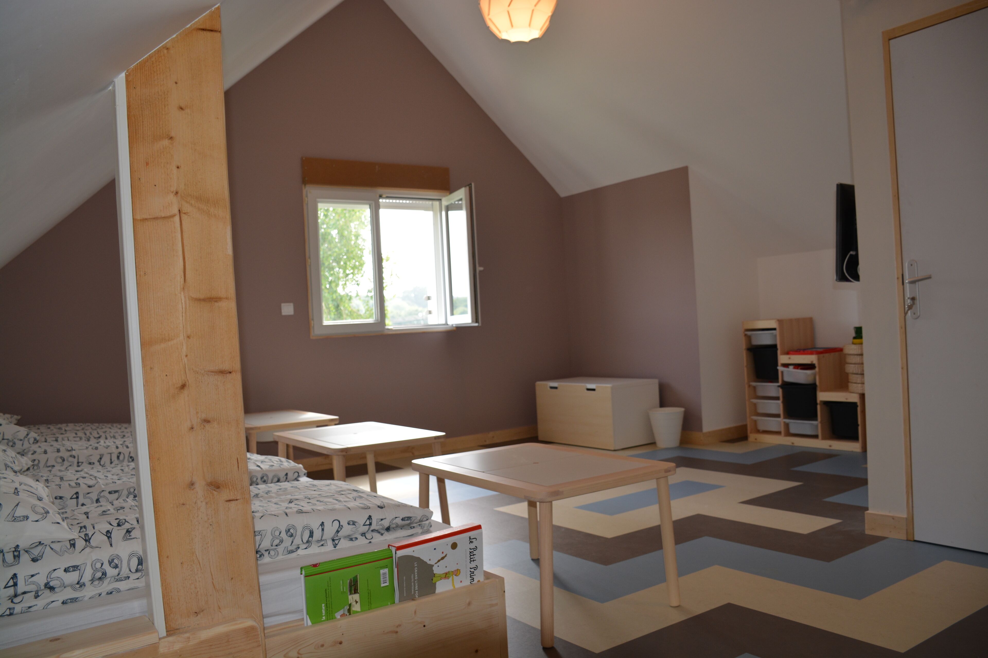 A room with a sloped ceiling, a window, a desk, a chair, and a storage unit.