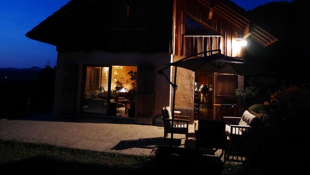 A two-story house with a large glass door entrance, outdoor seating area, and a well-lit interior. - Comfortable, quiet chalet (Les Adrets)