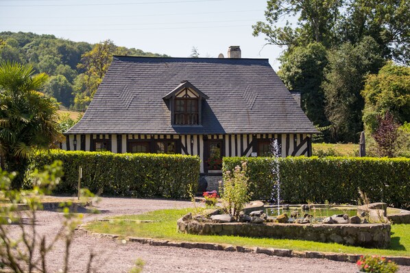 Une maison traditionnelle à colombages, dotée d’un toit à pignon, entourée de haies bien entretenues et d’un petit étang.