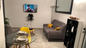 Ein kompakter Wohnbereich mit einem Glastisch, gelben Stühlen, einem grauen Sofa mit gelben Kissen, einem an der Wand montierten Flachbildfernseher und einer schwarzen Schrank mit Regalen.