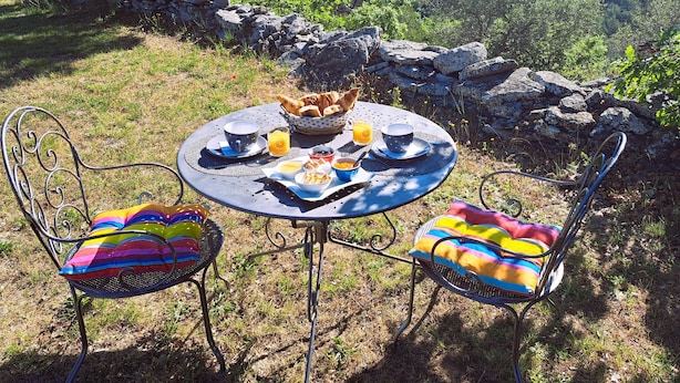 A round table with a stone wall backdrop, two metal chairs with colorful cushions, and a breakfast spread including bread, eggs, and orange juice. - Cante Grillet in Provence at the foot of the Luberon LACOSTE (84480) Chambre FLORY (LACOSTE)
