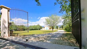 A gated entrance with a metal gate, a concrete pathway, a tree, and a building in the background. - Cante Grillet in Provence at the foot of the Luberon LACOSTE (84480) Chambre FLORY (LACOSTE)