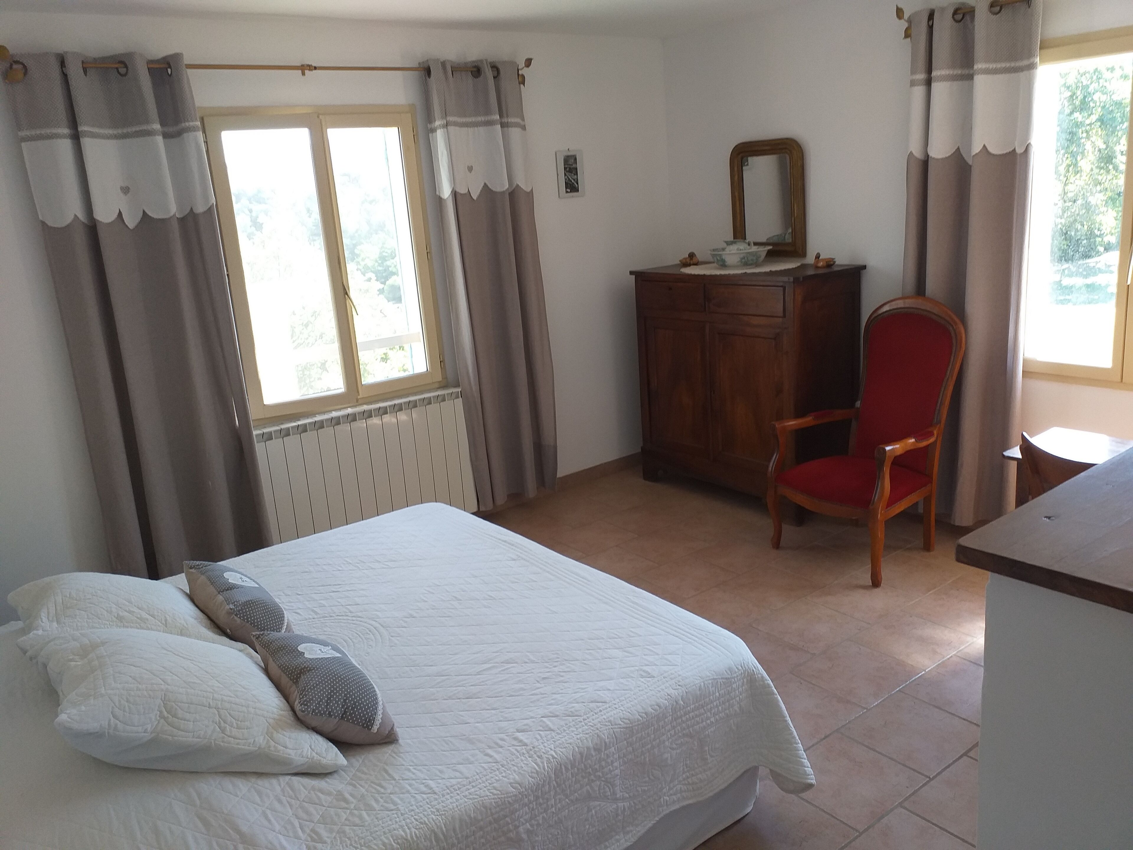 Ein Schlafzimmer mit einem Bett, einer Holzkommode, einem roten Stuhl und einem Fenster mit Vorhängen.
