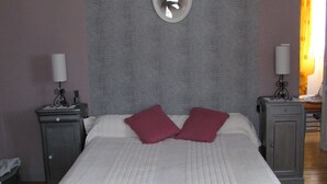 Ein ordentlich bezogenes Bett mit weißer Decke und zwei roten Kissen, ein kleiner Nachttisch mit Lampe und ein Spiegel an der Wand.