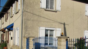 Ein Wohngebäude mit einer Stein-Fassade, mehreren Fenstern mit Fensterläden, einem blauen Metallzaun und einem Schotterweg.