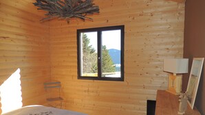 A wooden cabin bedroom with a bed, a window with a view of trees and mountains, a wooden dresser with a lamp, and a chair. - CERF apartment in new chalet with view of Matemale lake 12 beds (Les Angles)
