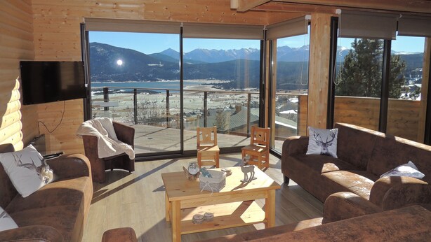 A wooden interior with a sofa, chairs, a table, and a TV. Large windows offer a view of mountains and a lake. - CERF apartment in new chalet with view of Matemale lake 12 beds (Les Angles)