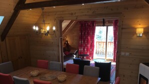 A wooden interior with a dining table, chairs, a chandelier, and a view to an outdoor area with a red curtain. - CHALET SABOT OF STANDING VENUS AT THE FOOT OF TRACKS (Fontcouverte-la-Toussuire)