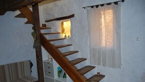A wooden staircase with a small window at the top, a white wall with textured plaster, and a white shelf unit with books and a basket. - Village house at the foot of Mont Ventoux (Bédoin)