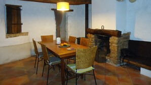 A rustic dining area with a wooden table, chairs, a hanging light, a stone fireplace, and a window with shutters. - Village house at the foot of Mont Ventoux (Bédoin)
