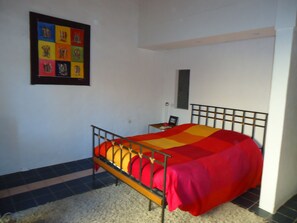 A bedroom with a metal bed frame, a colorful bedspread, a small bedside table with a lamp, and a framed picture on the wall.