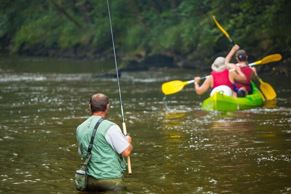 Un hombre pescando en un río, con un kayak al fondo.