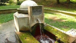 Una fuente de agua con un cartel de control de temperatura, rodeada por una cuenca de concreto y una zona verde.