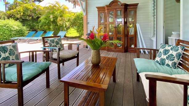 A wooden deck with lounge chairs and a table, a wooden cabinet, and a view of a pool and greenery.