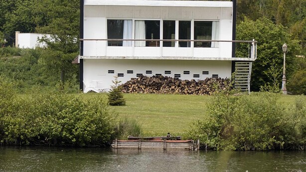 A modern two-story house with a white exterior, situated by a body of water, surrounded by lush greenery.