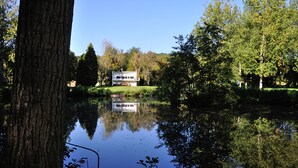 A lakeside view of a two-story building with a reflection in the water, surrounded by trees and greenery.