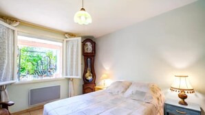 A bedroom with a bed, a nightstand, a lamp, a window with curtains, a radiator, and a wall-mounted clock. - MY LIFE Fayence Land of Flight (FAYENCE)