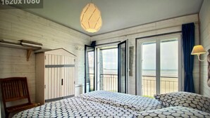 Ett rum med en säng, en träbänk, en hängande lampa och utsikt över stranden genom den öppna dörren.