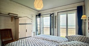 Una habitación con una cama, un banco de madera, una lámpara colgante y vista a la playa por la puerta abierta.