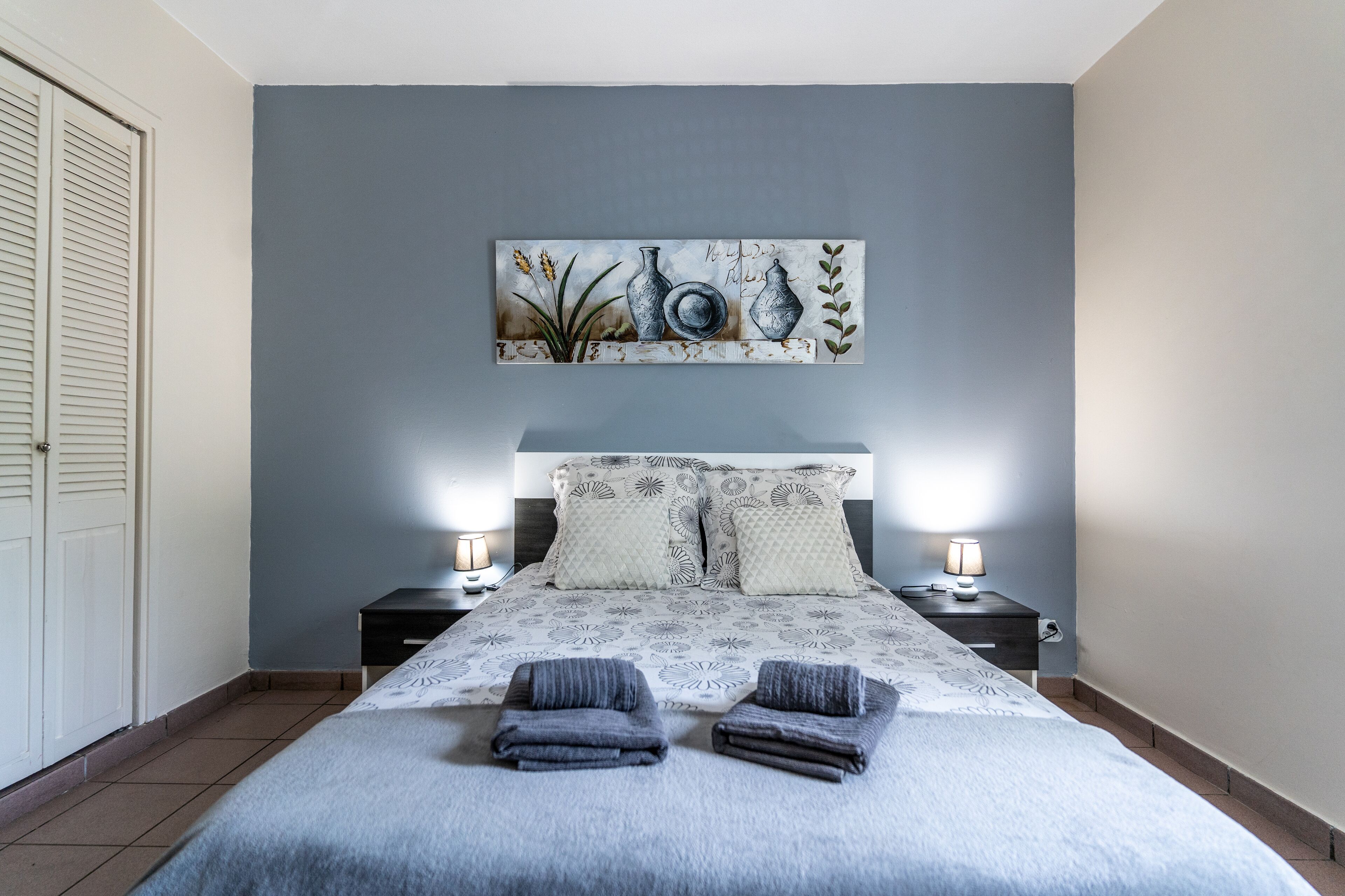 A bedroom with a bed, two bedside tables with lamps, a headboard with patterned pillows, and a wall-mounted painting.