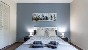 Une chambre à coucher avec un lit, deux tables de chevet avec des lampes, une tête de lit agrémentée d’oreillers à motifs et un tableau accroché au mur.