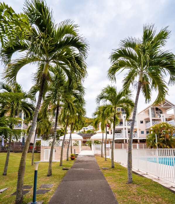 A pathway lined with palm trees leading to a building with multiple balconies and a pool area. - Le 222***, superb Duplex T4, residence with swimming pool, beach and restaurants 500m away (Le Diamant)
