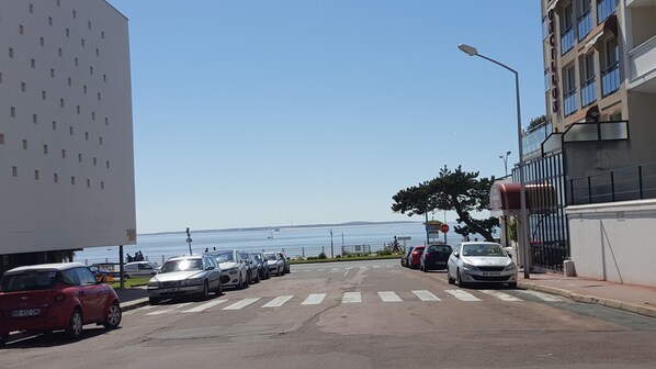 A street with parked cars, a pedestrian crossing, and a view of the sea.