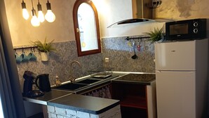 Une cuisine compacte dotée d’un îlot central intégré, d’un évier, d’une cuisinière et d’un réfrigérateur. Des luminaires sont suspendus au-dessus de l’îlot et une fenêtre se trouve au-dessus de l’évier.