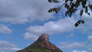 Un sentiero lastricato conduce a un colle con una cima rocciosa, circondato da vegetazione e un cielo sereno.