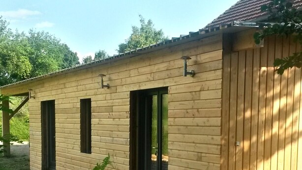 Een houten gebouw met drie deuren en ramen, omgeven door groen.