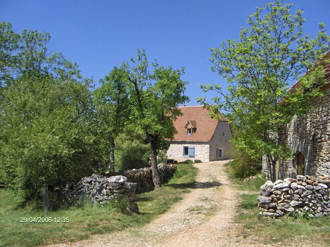 Une maison en pierre avec un toit rouge, entourée d’arbres et bordée d’un chemin de gravier.