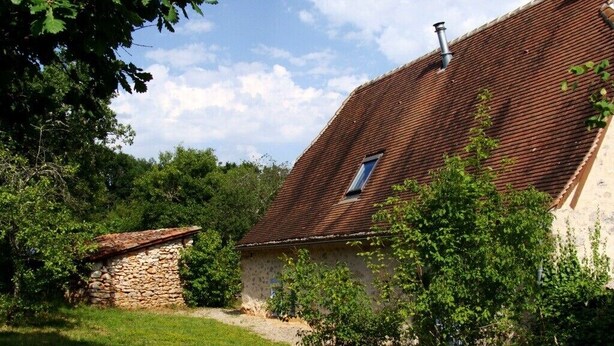 Een landelijk stenen gebouw met schuine kap, omgeven door groen en bomen.
