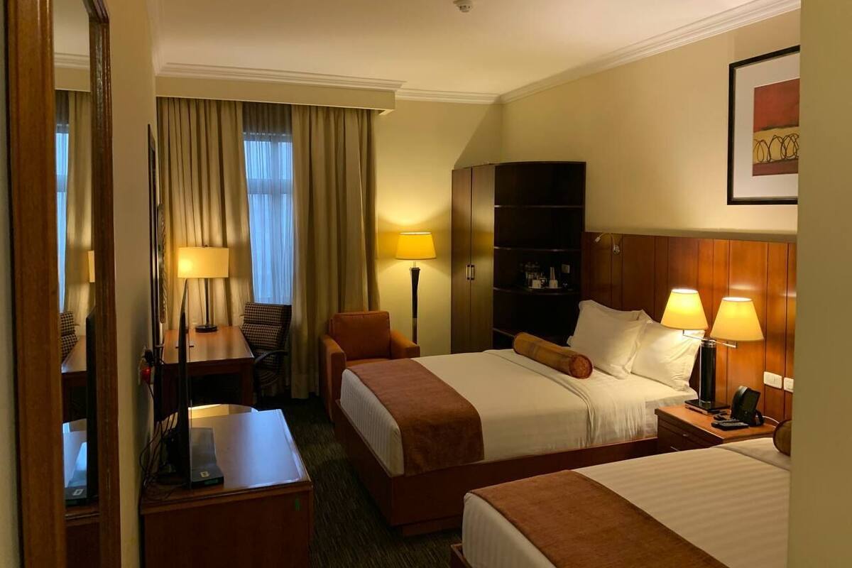 A hotel room with two beds, a desk, a chair, a wardrobe, and a lamp.