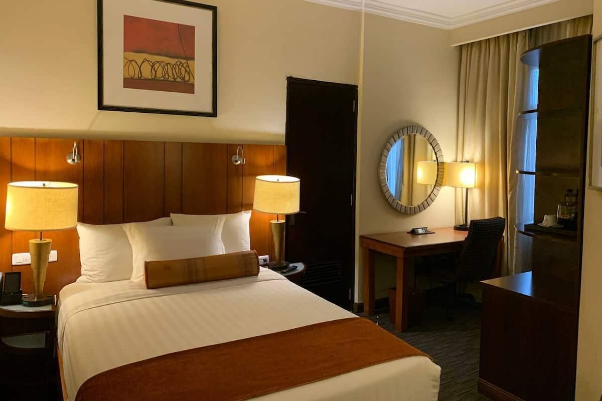 A hotel room with a large bed, two bedside lamps, a desk with a chair, a round mirror, and a framed picture on the wall.