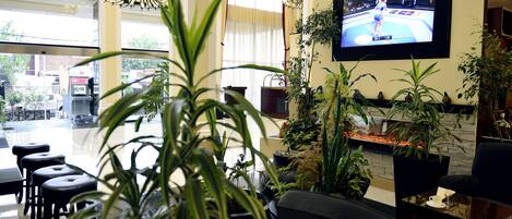 Un vestíbulo moderno con una televisión grande que muestra un evento deportivo, plantas en macetas y una zona de estar con taburetes.